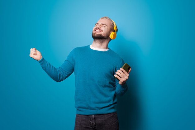 Hombre bailando con un teléfono está escuchando música con auriculares y sonríe en una pared azul en el estudio