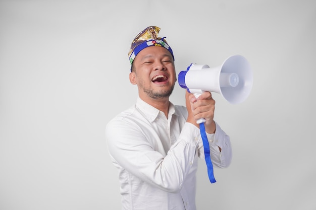 Foto hombre balinés enérgico con camisa blanca y tocado tradicional gritando en el megáfono mientras levanta el puño cerrado aislado por el fondo blanco