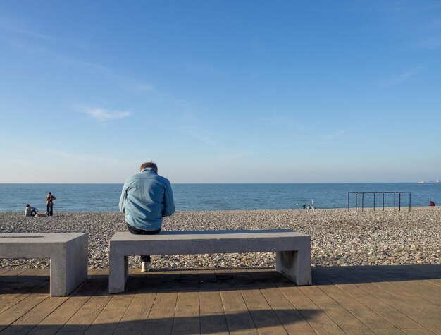 Hombre en un banco junto al mar Un hombre con un teléfono está sentado junto a una playa de guijarros Vacaciones en la playa Relajación en el mar Costa rocosa Playa de guijarros Barcos en el agua