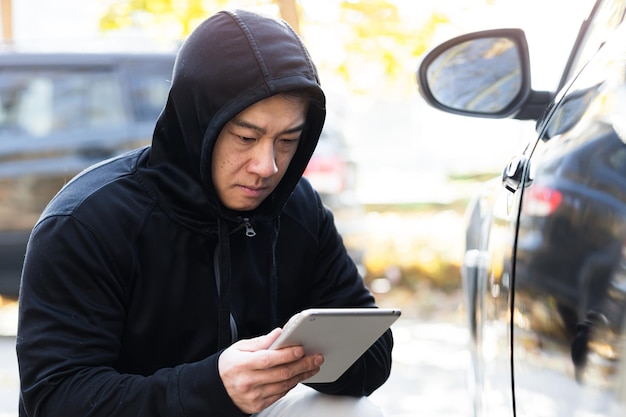 Hombre bandido ladrón ladrón de coches asiático utiliza una tableta para apagar la alarma del coche