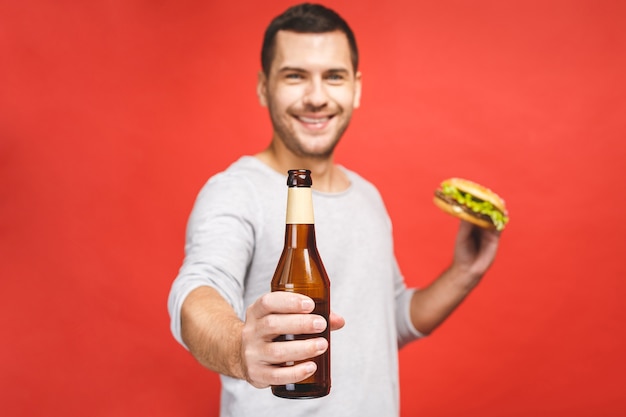 Hombre con barba aislado sobre fondo rojo sostiene una hamburguesa y una botella de cerveza, Retrato