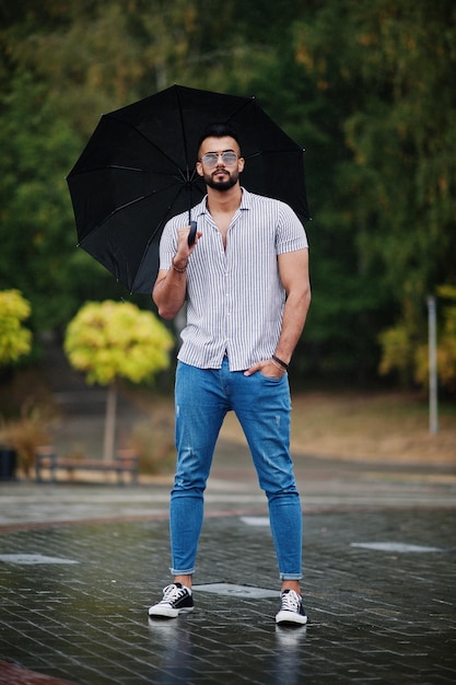 El hombre de barba árabe alto de moda usa jeans de camisa y gafas de sol con paraguas posados bajo la lluvia en la plaza del parque