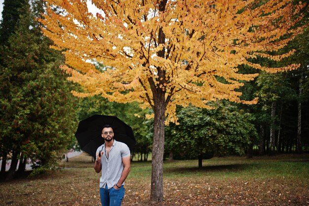 El hombre de barba árabe alto de moda usa pantalones vaqueros y gafas de sol posados en el parque de otoño con paraguas contra el árbol de hojas amarillas