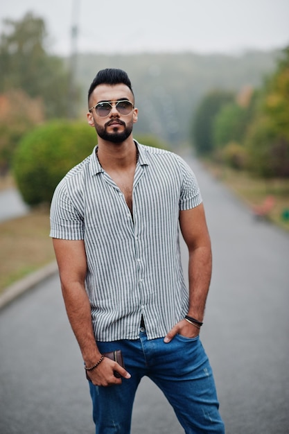 El hombre de barba árabe alto de moda usa pantalones vaqueros y gafas de sol posados en el parque y sostiene la billetera a mano