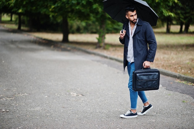 El hombre de barba árabe alto de moda viste un abrigo negro con paraguas y una bolsa que se presenta en el día del clima lluvioso