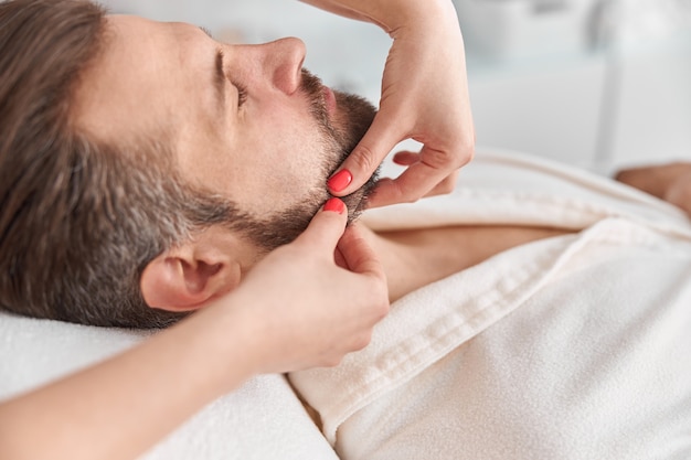 El hombre con barba está acostado boca arriba, recibiendo un masaje de estiramiento facial. Tratamiento de belleza de masaje facial. Concepto de bienestar, belleza y relajación.