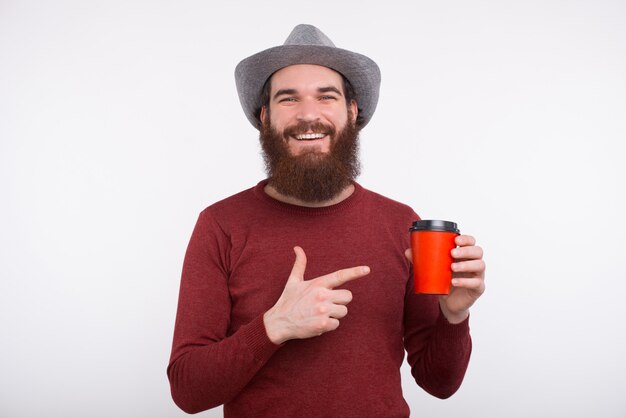 Un hombre con barba está apuntando a una taza con bebida caliente y sonriendo con un sombrero, cerca de la pared blanca.