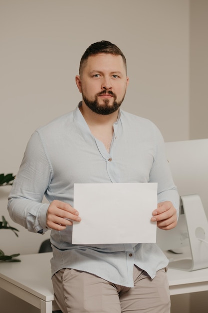 Un hombre con barba está sentado con una hoja vacía de papel blanco cerca de una computadora de escritorio de acero en casa