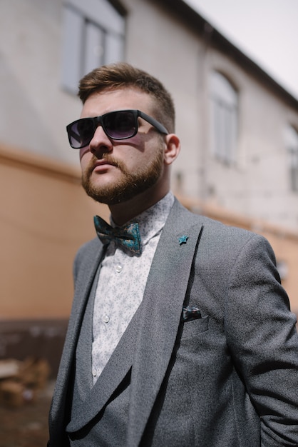 Un hombre con barba y gafas de sol con traje gris posa en la calle para anunciar la ropa de los hombres.