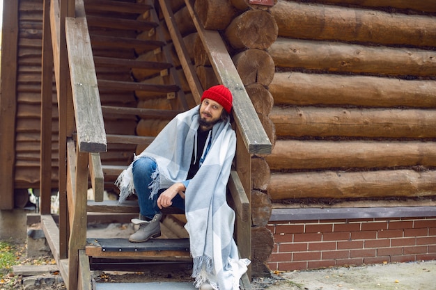 Hombre con barba hipster en un gorro de punto rojo y una chaqueta de mezclilla está sentado en las escaleras