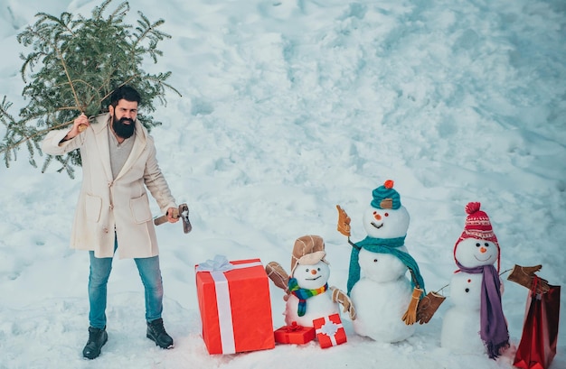 Hombre con barba lleva a casa un árbol de Navidad Emoción de invierno Feliz Navidad y Feliz Fiestas Joven leñador lleva abeto en el fondo de nieve blanca