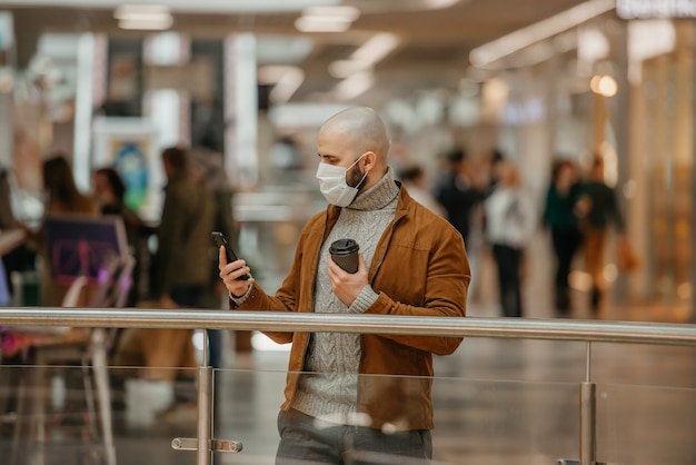 Un hombre con barba y mascarilla para evitar la propagación del coronavirus está usando un teléfono inteligente y sosteniendo una taza de café en el centro comercial. Un tipo calvo con una mascarilla quirúrgica mantiene la distancia social.