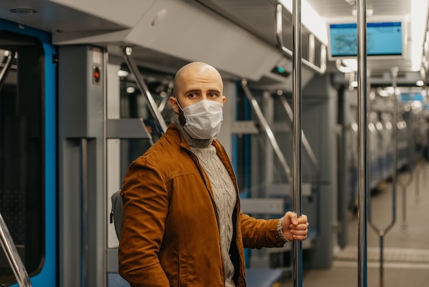 Un hombre con barba y una mascarilla médica para evitar la propagación del COVID-19 está de pie y agarrado del pasamanos en un vagón de metro. Un tipo calvo con una mascarilla quirúrgica mantiene distancia social en un tren.