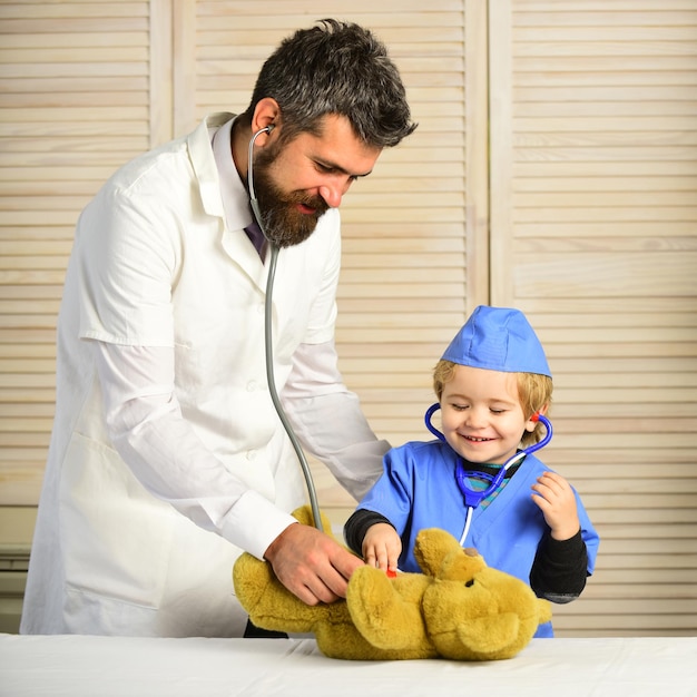 Hombre con barba y niño en batas médicas sostienen estetoscopio sobre fondo de madera Pediatra