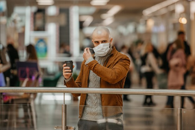 Un hombre con barba se pone una mascarilla médica mientras sostiene una taza de café en el centro comercial. Un tipo calvo mantiene distancia social.