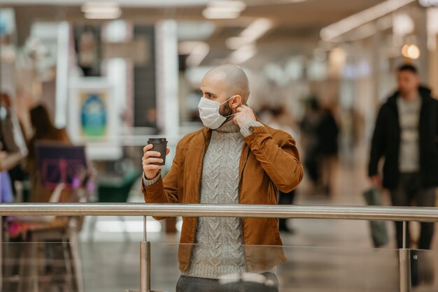 Un hombre con barba se pone una mascarilla médica mientras sostiene una taza de café marrón en el centro comercial. Un tipo calvo mantiene distancia social.