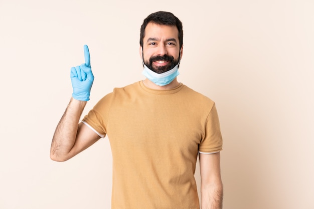 Hombre con barba protegiéndose del coronavirus con una máscara y guantes sobre una pared aislada apuntando una gran idea