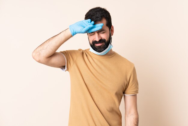 Hombre con barba protegiéndose del coronavirus con una máscara y guantes sobre pared aislada con expresión cansada y enferma