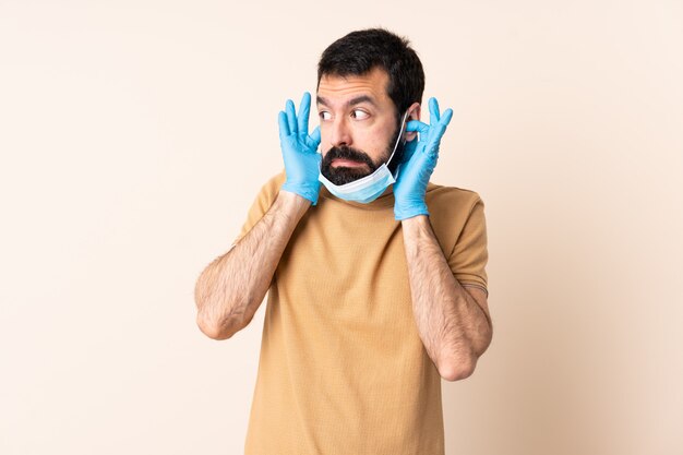 Hombre con barba protegiéndose del coronavirus con una máscara y guantes sobre pared aislada frustrado y cubriendo las orejas