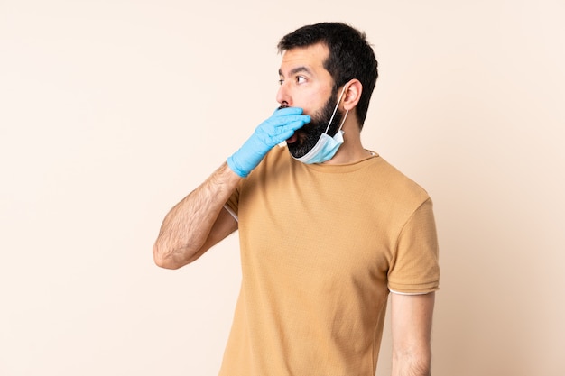 Hombre con barba protegiéndose del coronavirus con una máscara y guantes sobre pared aislada haciendo un gesto de sorpresa mientras mira a un lado