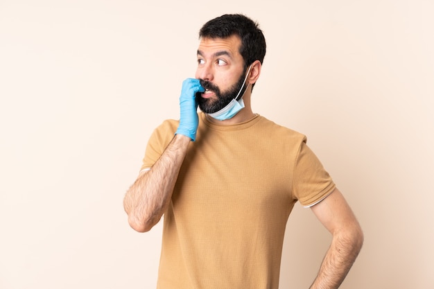 El hombre con barba que protege del coronavirus con una máscara y guantes sobre una pared aislada está un poco nervioso