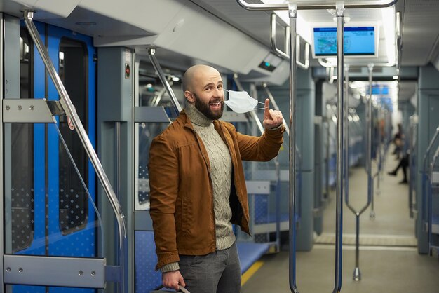 Foto un hombre con barba se quita una mascarilla médica y sonríe en un vagón de metro un tipo calvo