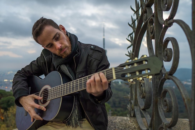 Hombre con barba roja tocando la guitarra sentado solo al lado de la valla