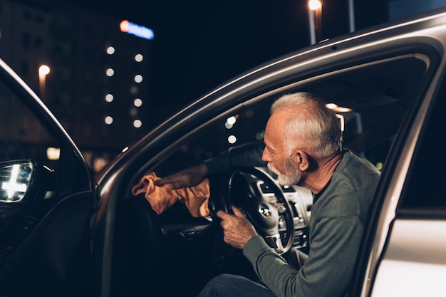 Hombre de barba Senior limpiando su coche con aspiradora por la noche en la estación de lavado de coches