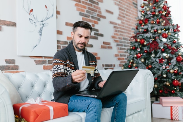 Un hombre con barba sentado en el sofá durante las vacaciones de Año Nuevo, nervioso y preocupado tratando de obtener un préstamo en línea, usando una computadora portátil y una tarjeta de crédito