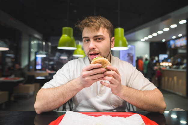 hombre con barba se sienta en un restaurante de comida rápida con una hamburguesa en sus manos