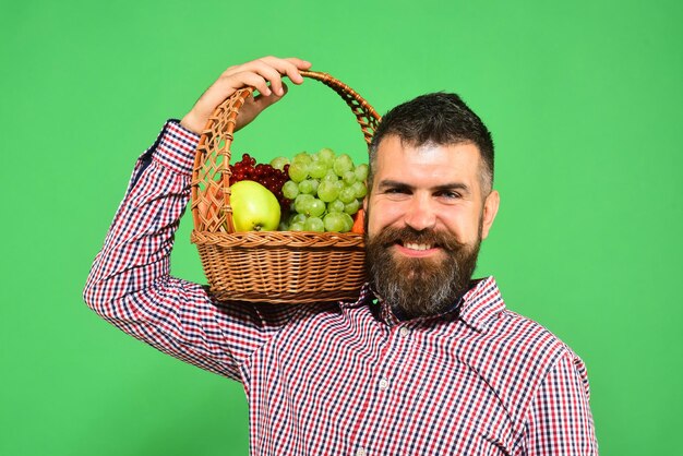 El hombre con barba sostiene una cesta con fruta en el hombro sobre fondo verde. Granjero con cara feliz presenta manzanas, uvas y arándanos. Guy tiene cosecha local. Concepto de agricultura y jardinería.