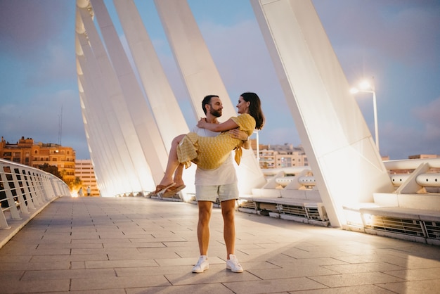 Un hombre con barba sostiene en sus brazos a su chica con un vestido amarillo en un puente blanco en Valencia. Un par de turistas en toda su longitud en una cita en la cálida noche.