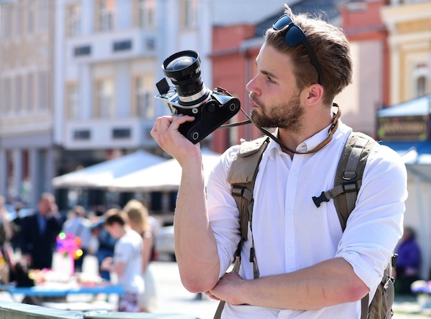 El hombre con barba tiene una cámara fotográfica en el fondo urbano. El turista toma una foto del paisaje urbano. Joven viajero o fotógrafo con cara curiosa va de turismo. Concepto de fotografía y turismo.