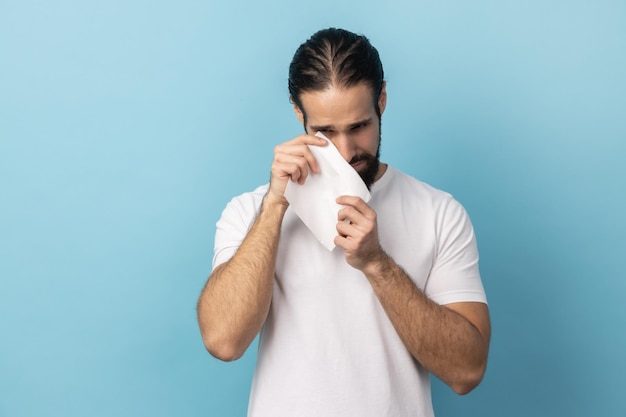 Hombre con barba usando camiseta de pie sosteniendo su cabeza hacia abajo y llorando con serios problemas