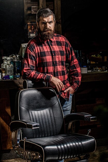 Foto hombre barbero en camisa de pie cerca de la silla en el salón de belleza hombre barbudo de pie en la barbería