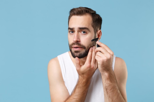 Hombre barbudo de 20 años con camisa blanca sostiene una maquinilla de afeitar de plástico desechable aislada en un retrato de estudio de fondo azul pastel. Concepto de procedimientos cosméticos para el cuidado de la piel. Simulacros de espacio de copia.