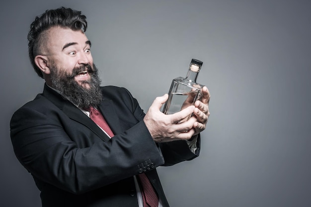 Hombre barbudo adulto feliz en traje mirando una botella de alcohol en la mano