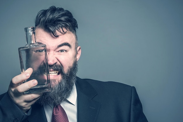 Hombre barbudo adulto malvado con traje mirando a través de una botella verde de alcohol en un fondo gris