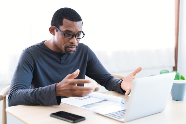 Hombre barbudo afroamericano confiado que usa anteojos trabaja en casa mientras habla de una reunión de videollamada a través de una computadora portátil en la oficina en casa Trabajo de educación remota desde el concepto de casa