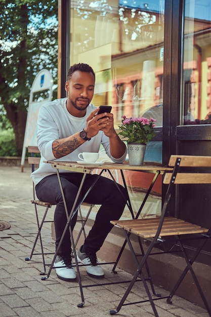 Hombre barbudo afroamericano de moda sentado cerca de una cafetería con una taza de café, usando un teléfono inteligente.