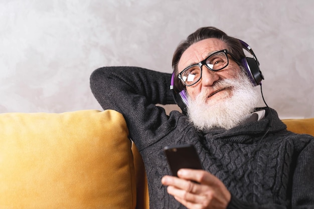 Hombre barbudo alegre de alto rango con anteojos que usan un suéter gris que se relaja con el teléfono inteligente mientras se sienta con auriculares en un sofá amarillo en su sala de estar luminosa concepto de comunicación de tecnología moderna