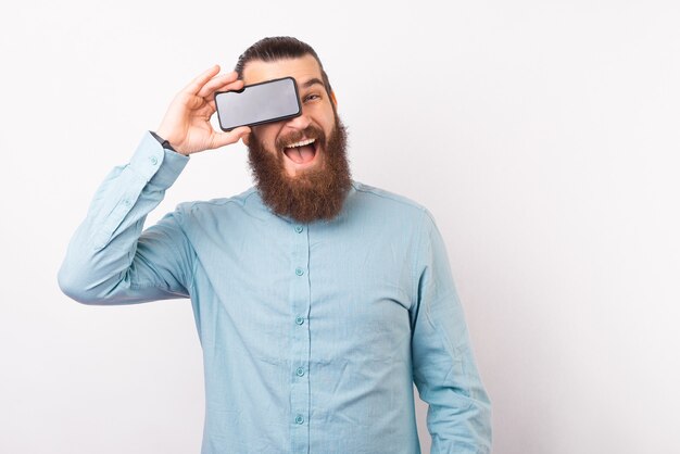 Hombre barbudo alegre en casual sosteniendo smartphone sobre sus ojos y sonriendo