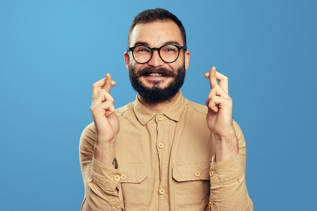 Hombre barbudo alegre mantiene los dedos cruzados hace que el deseo desee contra el azul