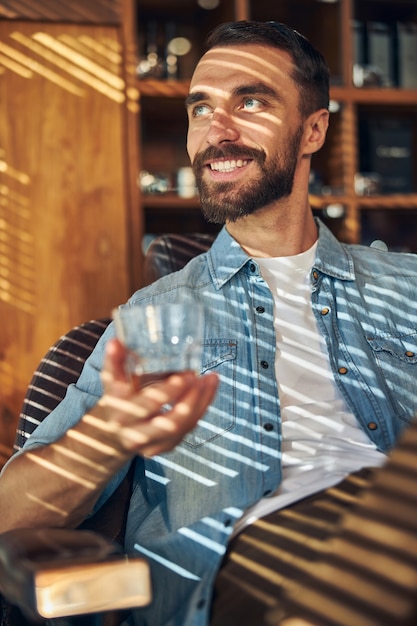 Un hombre barbudo alegre que sostiene una copa de whisky mientras está sentado en un sillón en la barbería