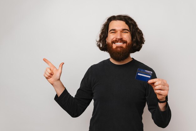 El hombre barbudo alegre señala a un lado y sostiene una tarjeta de crédito azul