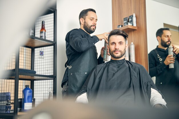 Hombre barbudo alegre sentado en un cómodo sillón y mirando hacia adelante mientras escucha a su peluquero