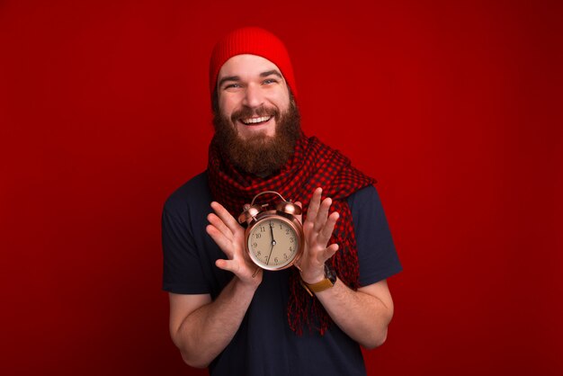 Hombre barbudo alegre, con sombrero rojo y bufanda, sosteniendo un reloj vintage, de pie sobre el espacio rojo