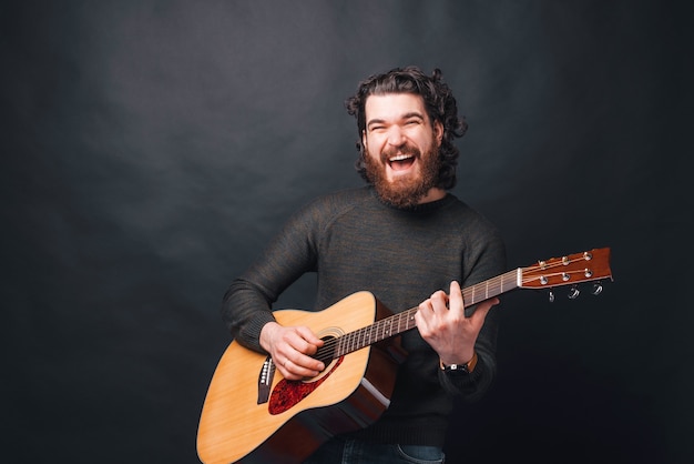 Hombre barbudo alegre tocando la guitarra acústica cerca de la pared oscura