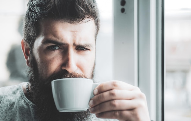 Hombre barbudo de aspecto gruñón bebiendo una taza de café por la mañana cerca de la ventana hipster hombre tomando una taza de amer