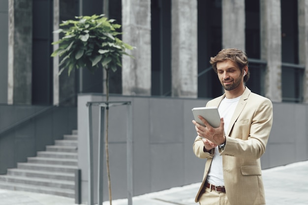 Hombre barbudo atractivo en traje formal sosteniendo tableta digital mientras está de pie cerca del edificio de oficinas